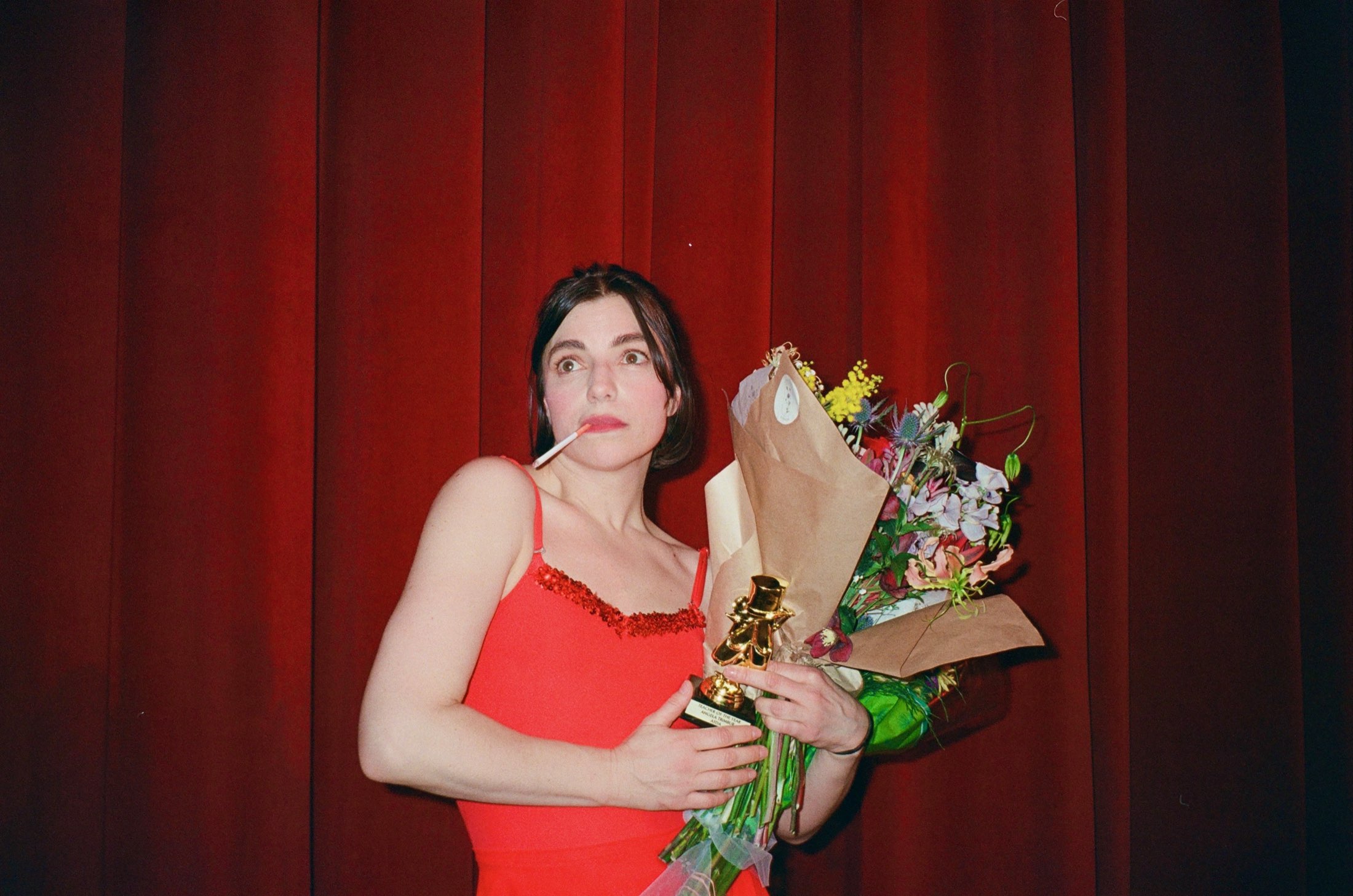 Angela Trimbur in a red leotard against red stage curtains, a lollipop in her mouth, holding a gold Teacher of the Year trophy and a bouquet